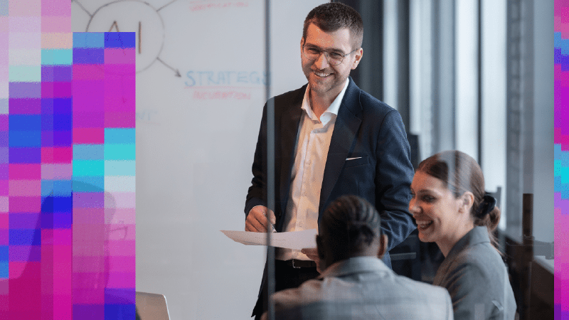 Smiling man in a suit presenting to two colleagues at a table by a whiteboard sketching an AI strategy