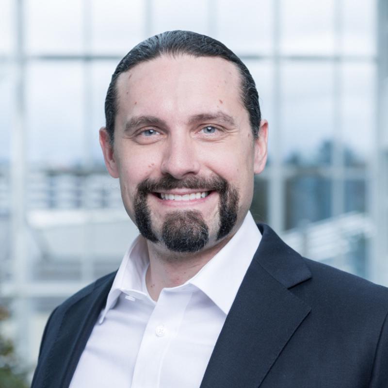 Portrait of Romano Roth smiling in a dark suit and open white shirt in front of a bright office window